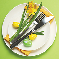 A white plate with fork and knife, flowers, a yellow napkin and yellow and green easter eggs